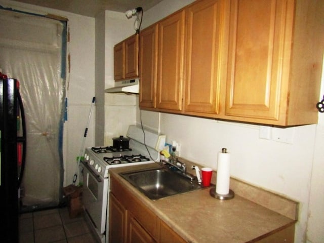 kitchen with a sink, under cabinet range hood, white range with gas stovetop, brown cabinetry, and freestanding refrigerator