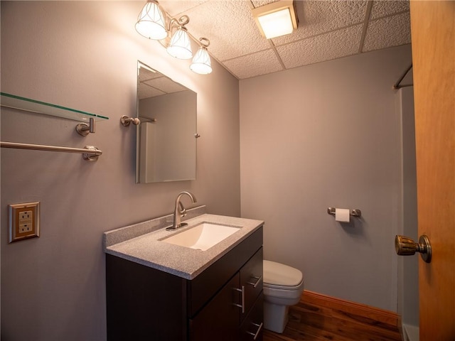 bathroom featuring a paneled ceiling, toilet, vanity, wood finished floors, and baseboards