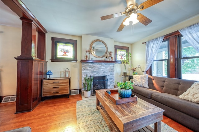 living room featuring ceiling fan, a stone fireplace, light wood-type flooring, and decorative columns