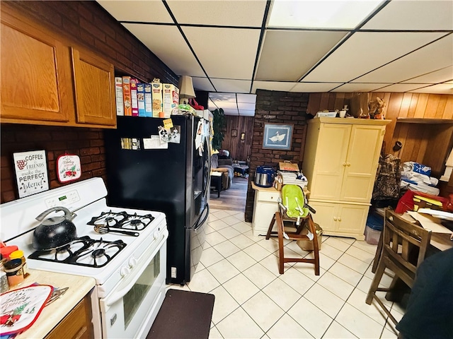 kitchen with a paneled ceiling, wood walls, light tile patterned floors, white gas stove, and brick wall