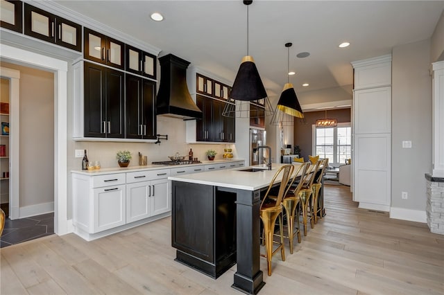 kitchen with custom exhaust hood, hanging light fixtures, sink, a center island with sink, and light hardwood / wood-style flooring