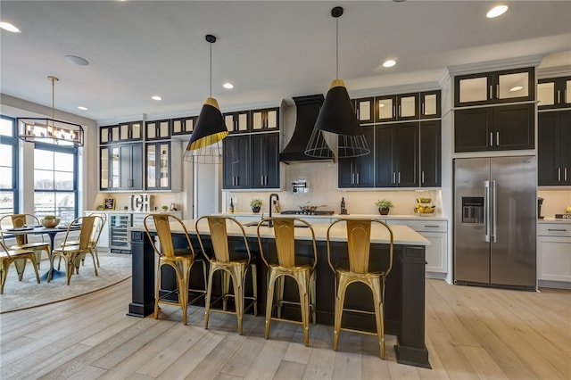 kitchen with custom range hood, light hardwood / wood-style floors, a center island with sink, decorative light fixtures, and high end refrigerator