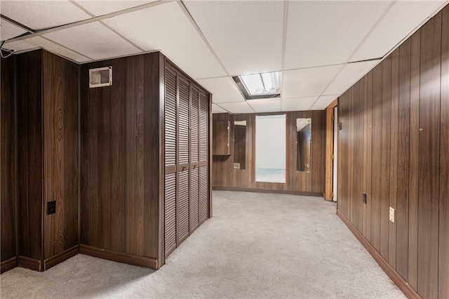 hallway with a paneled ceiling, wood walls, and light colored carpet