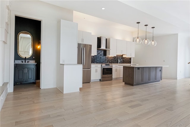 kitchen with wall chimney range hood, white cabinets, light hardwood / wood-style flooring, stainless steel appliances, and decorative light fixtures