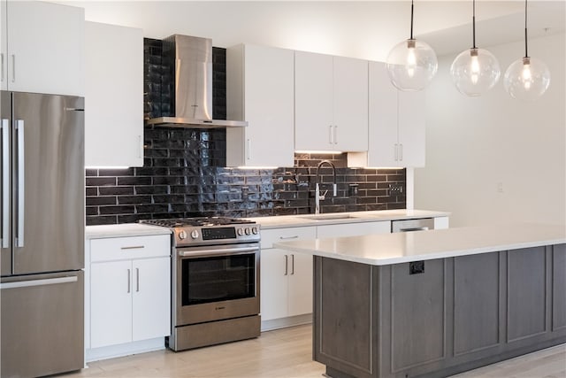 kitchen featuring wall chimney range hood, hanging light fixtures, stainless steel appliances, sink, and white cabinetry