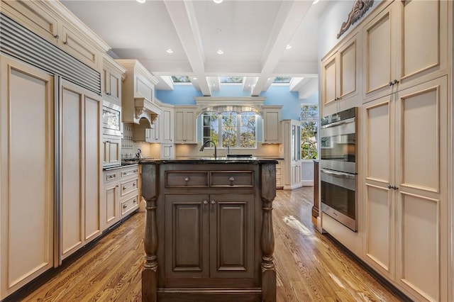 kitchen featuring light wood-type flooring, dark stone counters, cream cabinets, built in appliances, and beamed ceiling