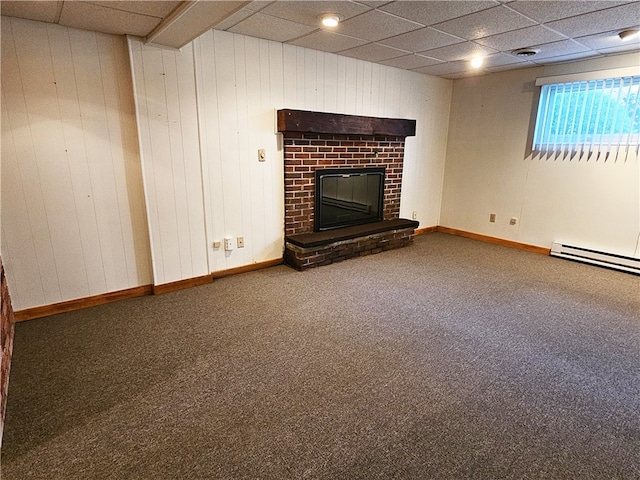 basement featuring baseboard heating, wooden walls, a drop ceiling, a brick fireplace, and carpet floors