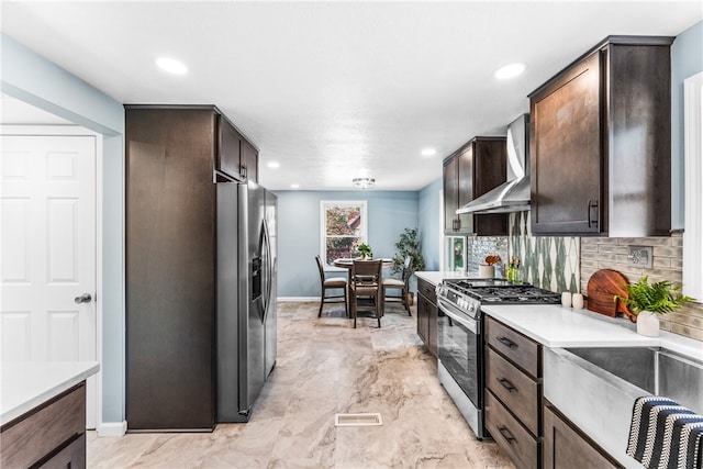 kitchen with wall chimney range hood, dark brown cabinets, tasteful backsplash, and appliances with stainless steel finishes