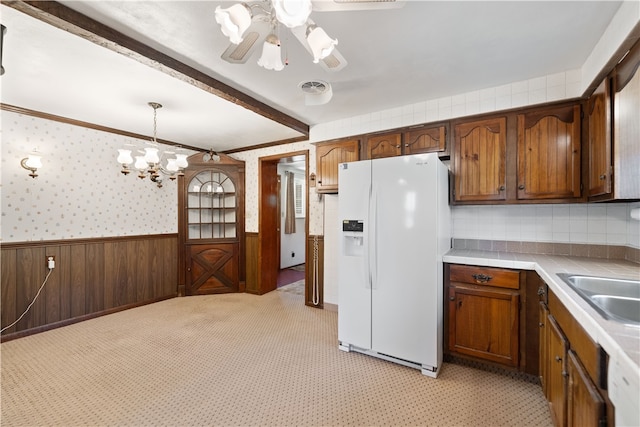 kitchen with white fridge with ice dispenser, wooden walls, sink, decorative light fixtures, and ceiling fan with notable chandelier