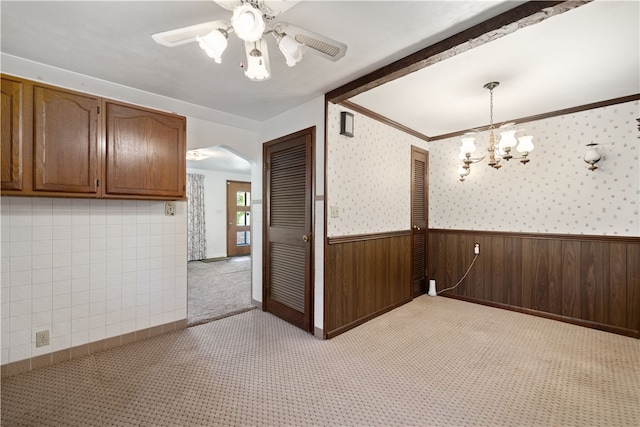 carpeted spare room featuring ornamental molding, ceiling fan with notable chandelier, and wood walls