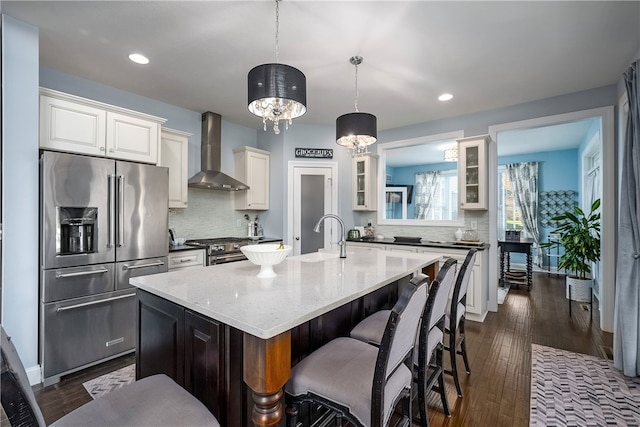 kitchen with wall chimney range hood, a center island with sink, sink, decorative light fixtures, and high end appliances