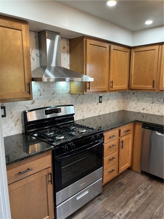 kitchen featuring wall chimney range hood, decorative backsplash, dark hardwood / wood-style flooring, dark stone counters, and appliances with stainless steel finishes