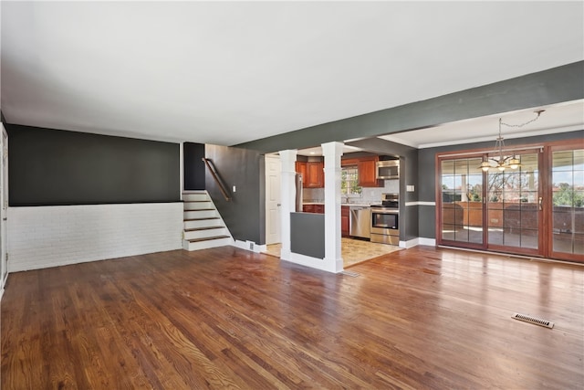 unfurnished living room featuring ornate columns, a notable chandelier, light wood-type flooring, and brick wall