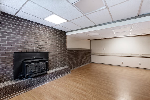 basement with a paneled ceiling, brick wall, a wood stove, and light hardwood / wood-style flooring