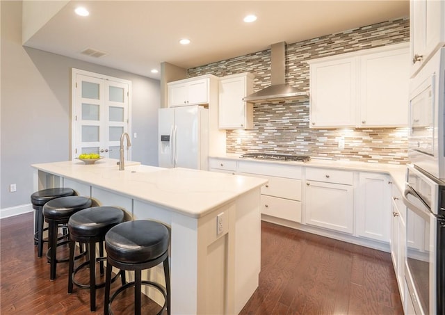 kitchen with backsplash, wall chimney range hood, an island with sink, white cabinetry, and stainless steel appliances