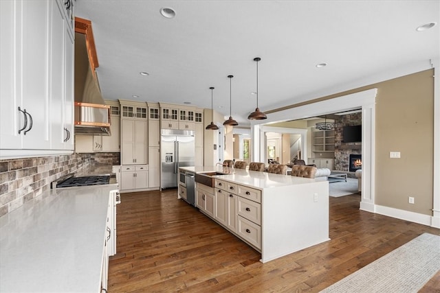 kitchen featuring appliances with stainless steel finishes, an island with sink, a fireplace, decorative light fixtures, and dark wood-type flooring