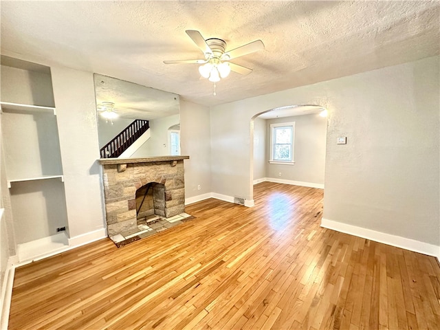 unfurnished living room with light hardwood / wood-style floors, a stone fireplace, a textured ceiling, and ceiling fan