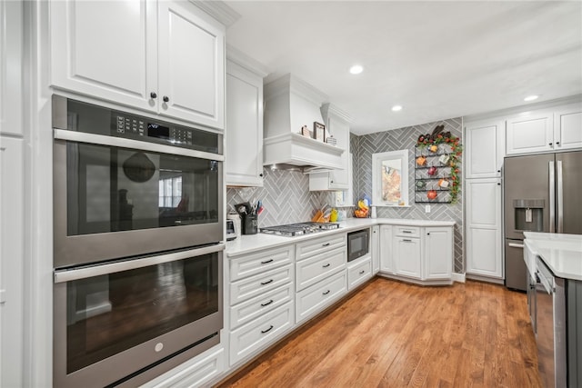 kitchen with appliances with stainless steel finishes, white cabinetry, premium range hood, light hardwood / wood-style floors, and tasteful backsplash