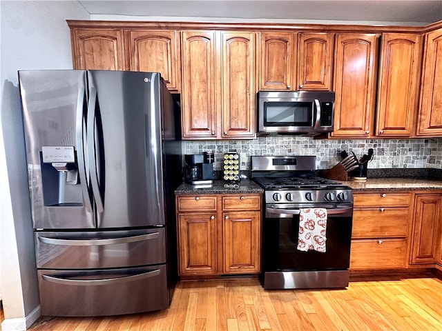 kitchen with dark stone countertops, stainless steel appliances, light wood-type flooring, and backsplash