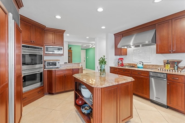 kitchen with light stone countertops, wall chimney range hood, a center island, stainless steel appliances, and decorative backsplash