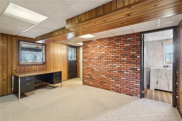 carpeted home office with a healthy amount of sunlight, a paneled ceiling, wood walls, and brick wall