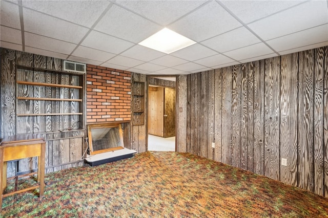 unfurnished living room with wooden walls, a brick fireplace, a paneled ceiling, and carpet floors