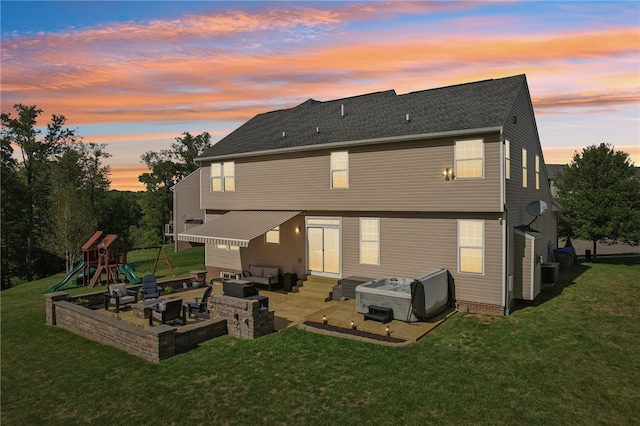 back house at dusk featuring a hot tub, a yard, a patio area, and a playground