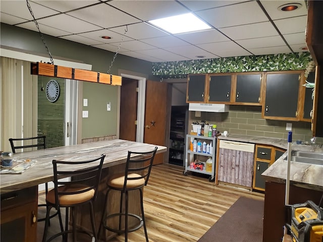 kitchen featuring a paneled ceiling, light wood-type flooring, white dishwasher, decorative backsplash, and a kitchen breakfast bar