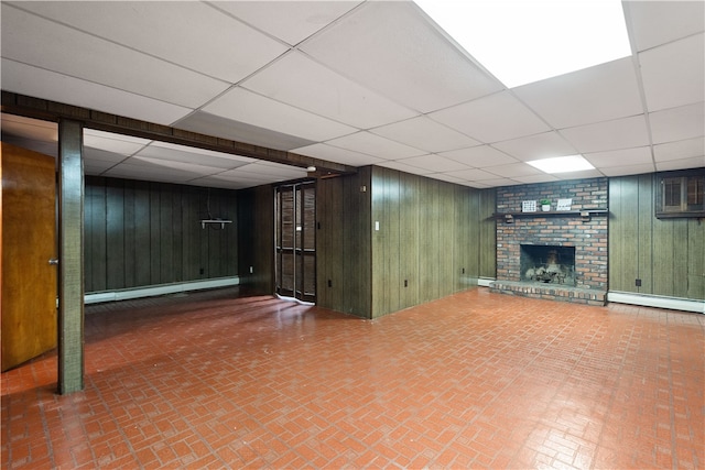 basement with a drop ceiling, a brick fireplace, baseboard heating, and wooden walls