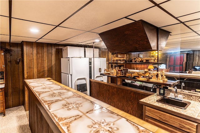 kitchen with sink, a drop ceiling, wooden walls, white fridge, and carpet floors