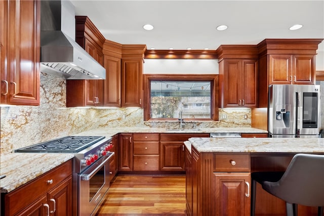 kitchen featuring wall chimney range hood, appliances with stainless steel finishes, light wood-type flooring, light stone countertops, and sink