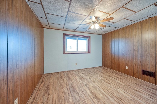 empty room with a drop ceiling, light hardwood / wood-style floors, ceiling fan, and wood walls