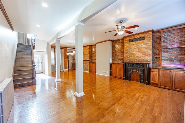 unfurnished living room featuring ceiling fan with notable chandelier, decorative columns, ornamental molding, light wood-type flooring, and brick wall