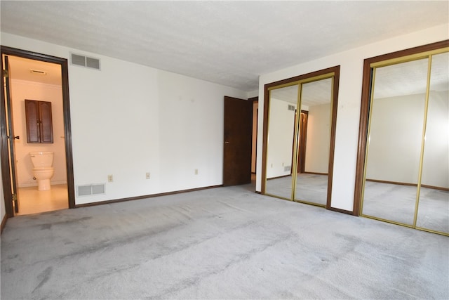 unfurnished bedroom featuring ensuite bath, a textured ceiling, light colored carpet, and two closets