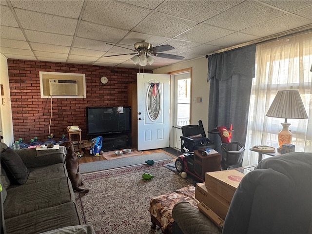 living room featuring a wall unit AC, hardwood / wood-style floors, a paneled ceiling, ceiling fan, and brick wall