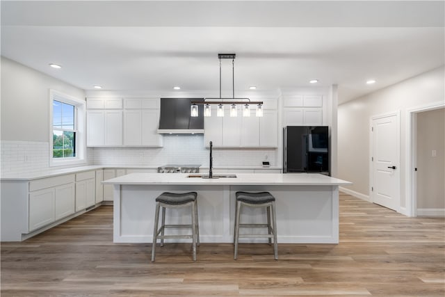 kitchen with a kitchen island with sink, custom range hood, black refrigerator, sink, and light hardwood / wood-style flooring