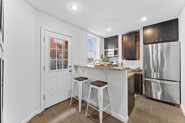 kitchen featuring stainless steel appliances, dark brown cabinetry, a breakfast bar, light stone counters, and kitchen peninsula