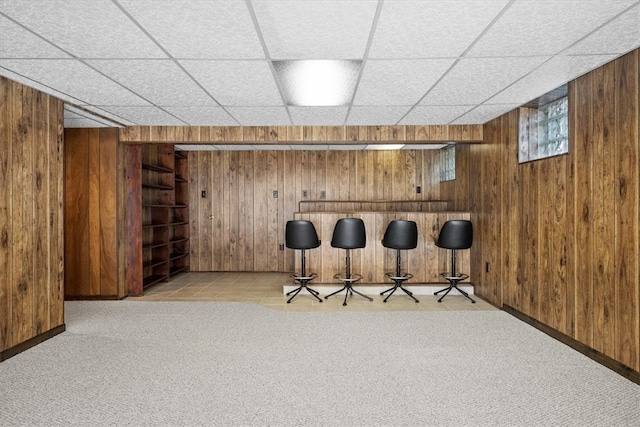 bar with a drop ceiling, light colored carpet, and wood walls