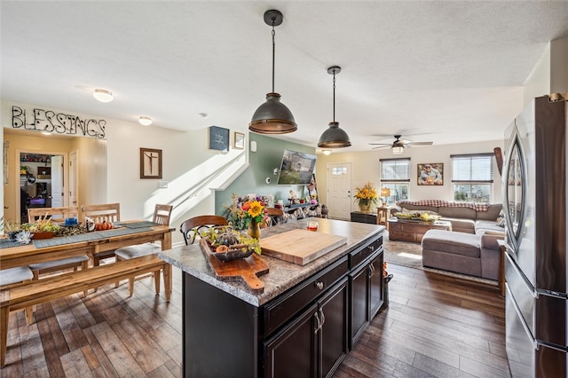 kitchen with dark wood-type flooring, hanging light fixtures, ceiling fan, a kitchen island, and stainless steel refrigerator