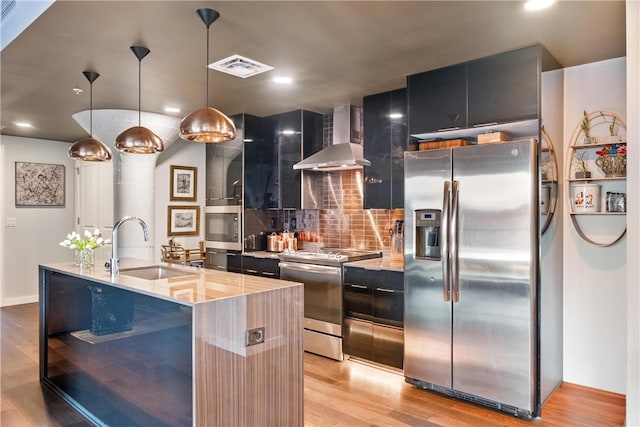 kitchen with sink, stainless steel appliances, wall chimney range hood, an island with sink, and decorative light fixtures