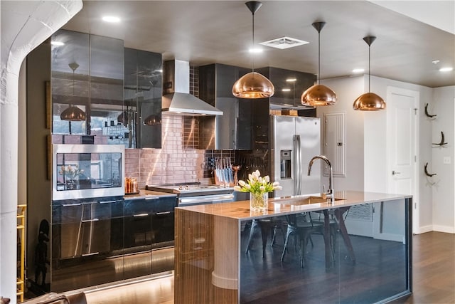 kitchen featuring stainless steel appliances, wall chimney range hood, wood-type flooring, decorative light fixtures, and a center island with sink