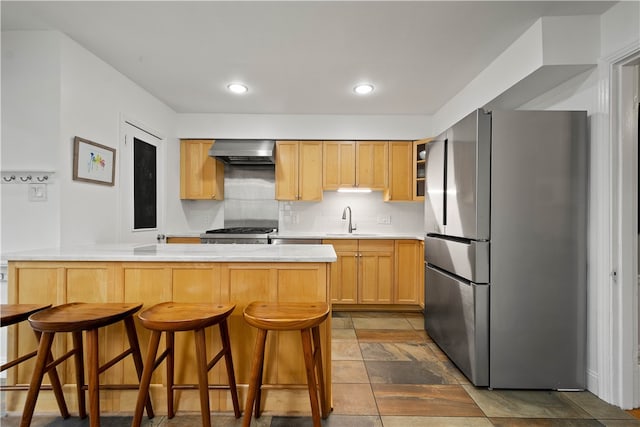 kitchen with appliances with stainless steel finishes, backsplash, a kitchen breakfast bar, wall chimney range hood, and light brown cabinets