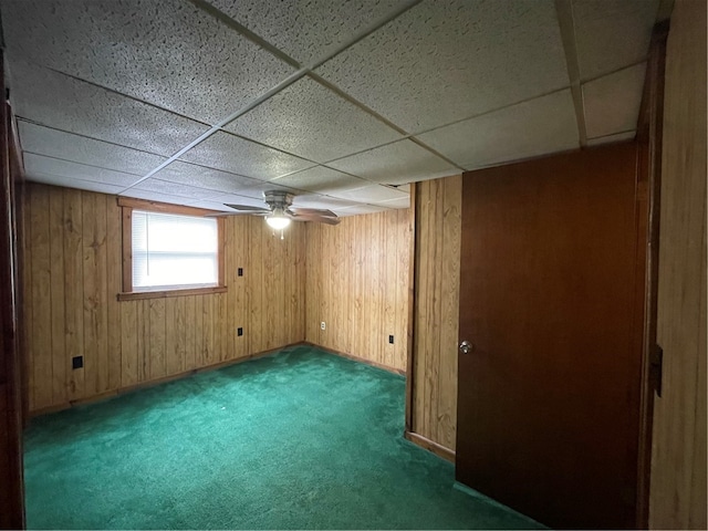 basement featuring wooden walls, a drop ceiling, ceiling fan, and dark colored carpet