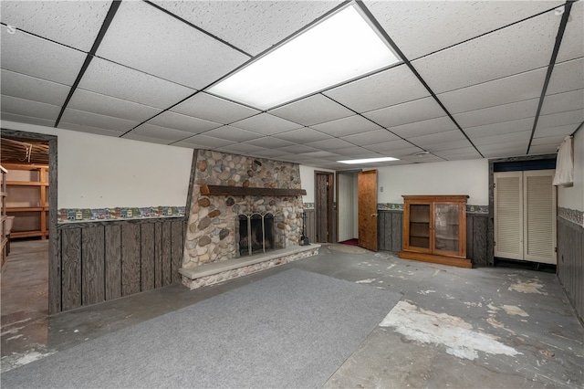 unfurnished living room featuring a paneled ceiling, a stone fireplace, concrete flooring, and wooden walls