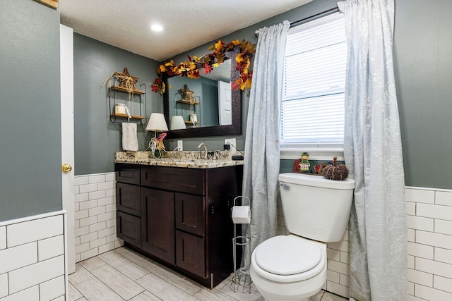 bathroom with a textured ceiling, vanity, toilet, and tile walls