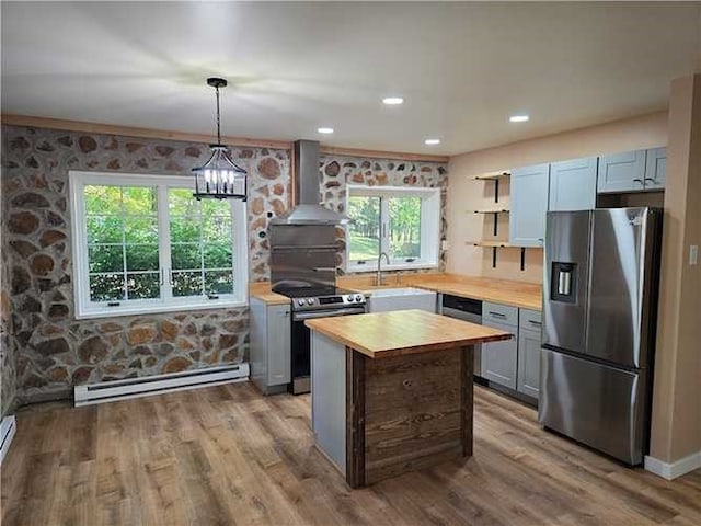 kitchen with a baseboard radiator, wall chimney range hood, light hardwood / wood-style floors, decorative light fixtures, and appliances with stainless steel finishes