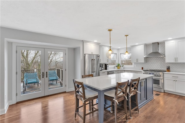 kitchen featuring french doors, wall chimney range hood, hanging light fixtures, appliances with stainless steel finishes, and wood-type flooring