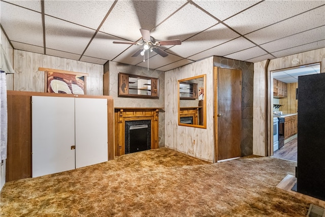 unfurnished living room with ceiling fan, wood walls, a drop ceiling, and carpet floors