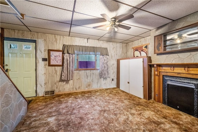 unfurnished living room with wood walls, a drop ceiling, ceiling fan, and carpet floors