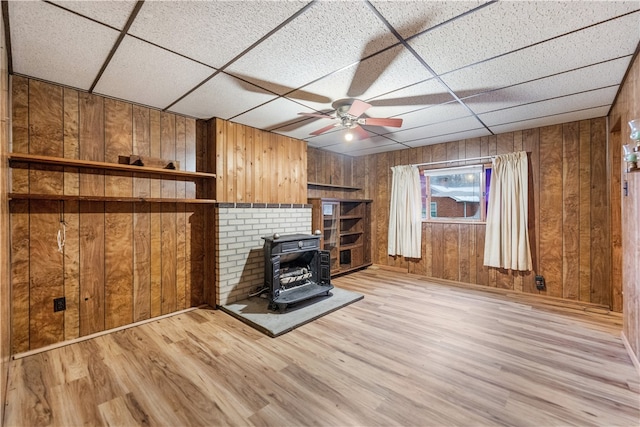 unfurnished living room with a paneled ceiling, ceiling fan, a wood stove, and wooden walls